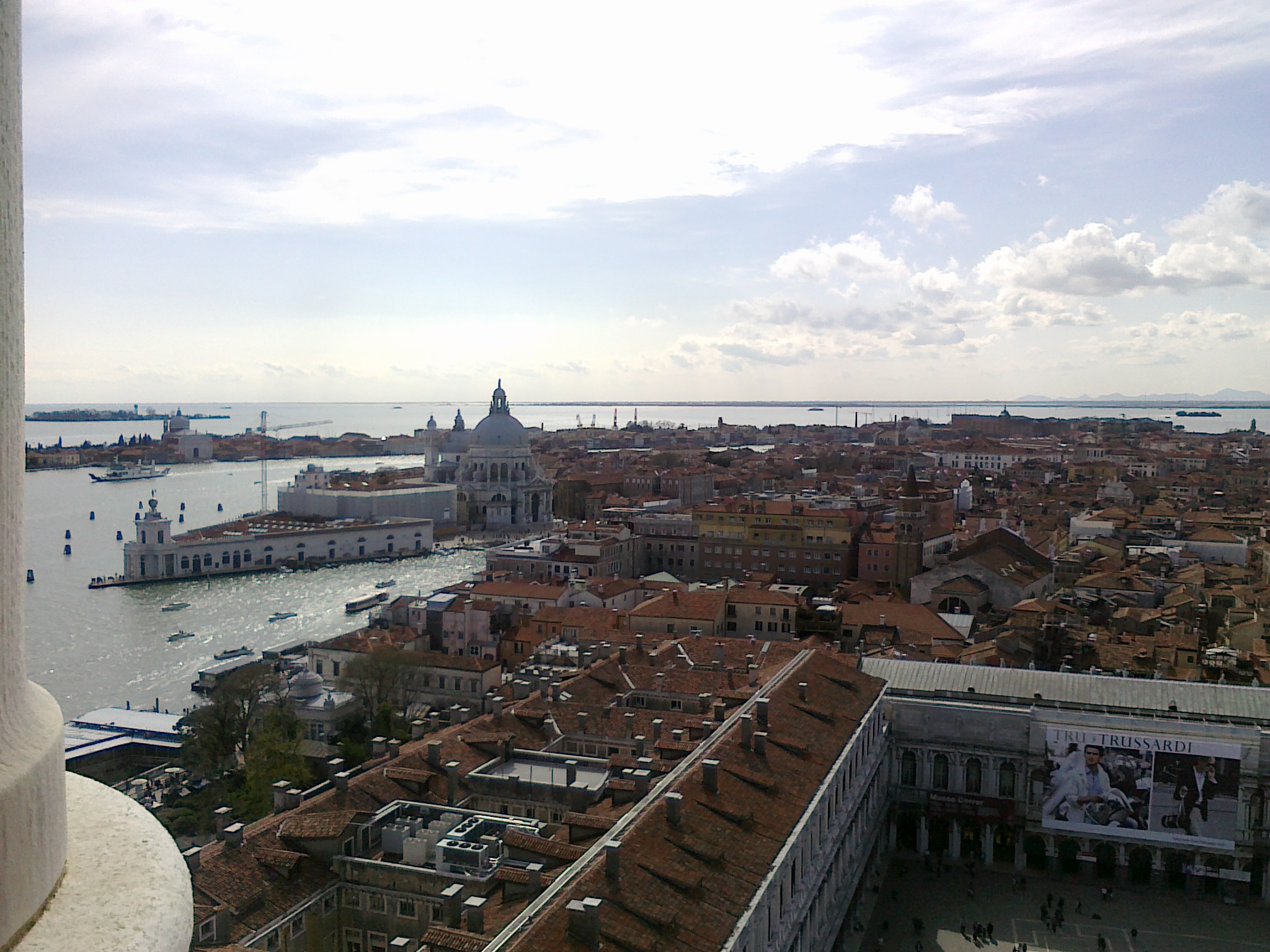 An aerial view of Venice's cityscape with numerous red rooftops and a wide canal and the sea in the background.